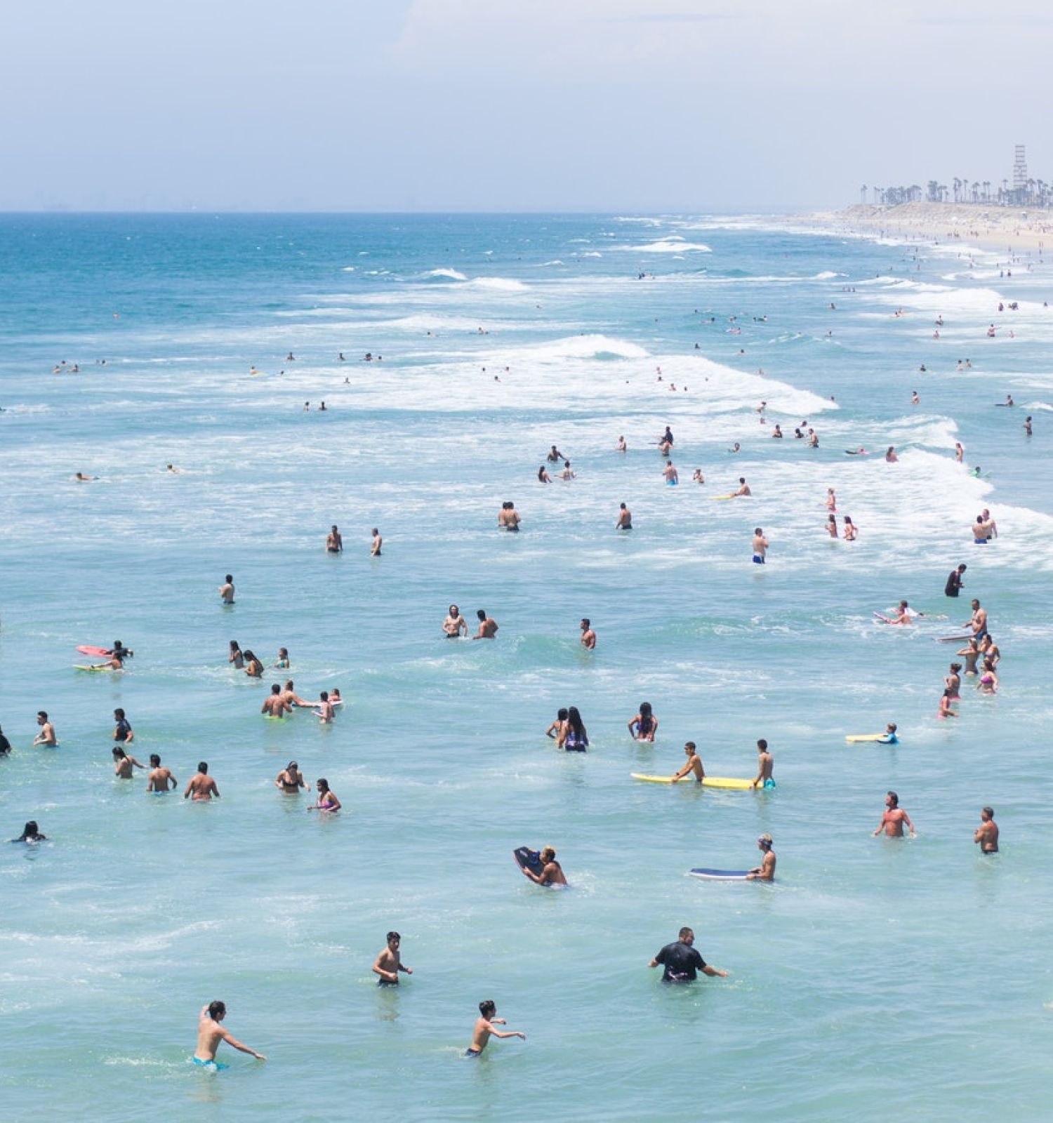 Many people are enjoying the ocean water at a crowded beach with waves crashing, some swimming while others are surfing or playing.