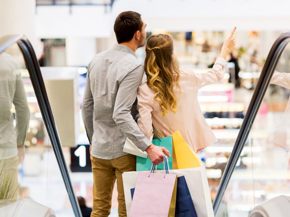 A couple stands on an escalator in a shopping mall, holding shopping bags, with the woman pointing towards something in the distance.