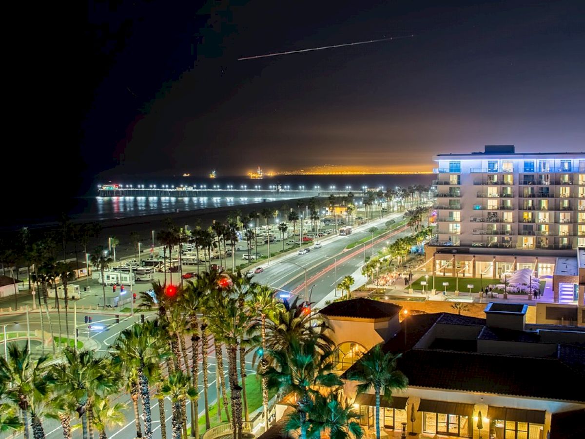 A nighttime coastal city view with a well-lit road, palm trees, modern buildings, and a pier extending into the illuminated ocean.