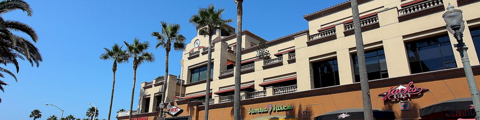 A street scene with palm trees, shops, and outdoor seating on a sunny day. People are walking, and cars are parked along the street.