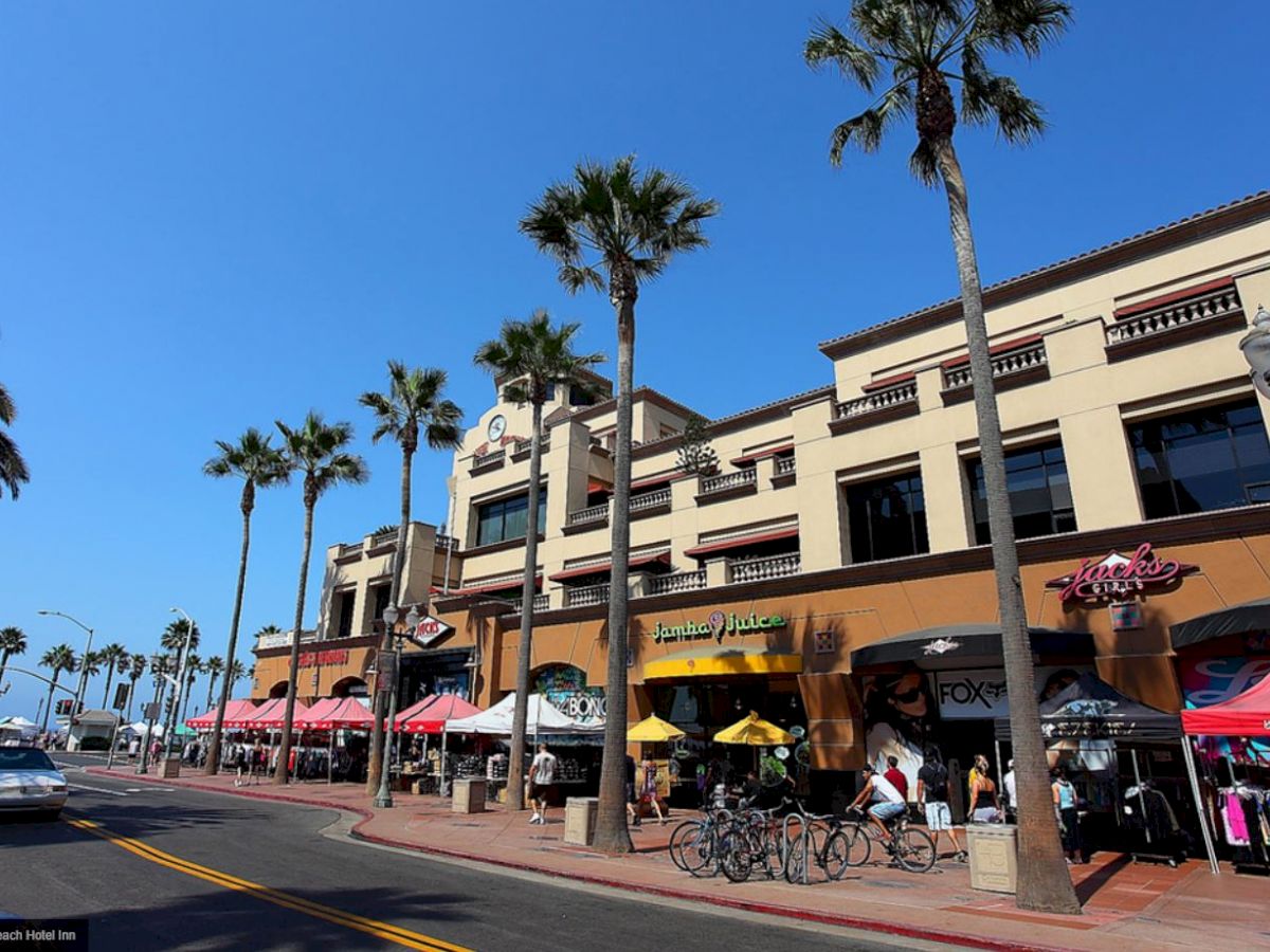 A bustling street scene with palm trees, storefronts, parked bicycles, and people enjoying a sunny day in front of a shopping complex.