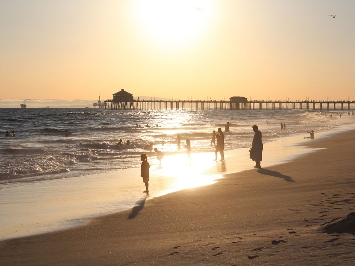 People are enjoying a sunset at the beach, with a long pier in the background and waves gently hitting the shore, creating a peaceful scene.