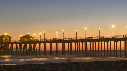A scenic pier extends over the ocean at sunset, with lights illuminating the surrounding buildings and casting a reflection on the water.