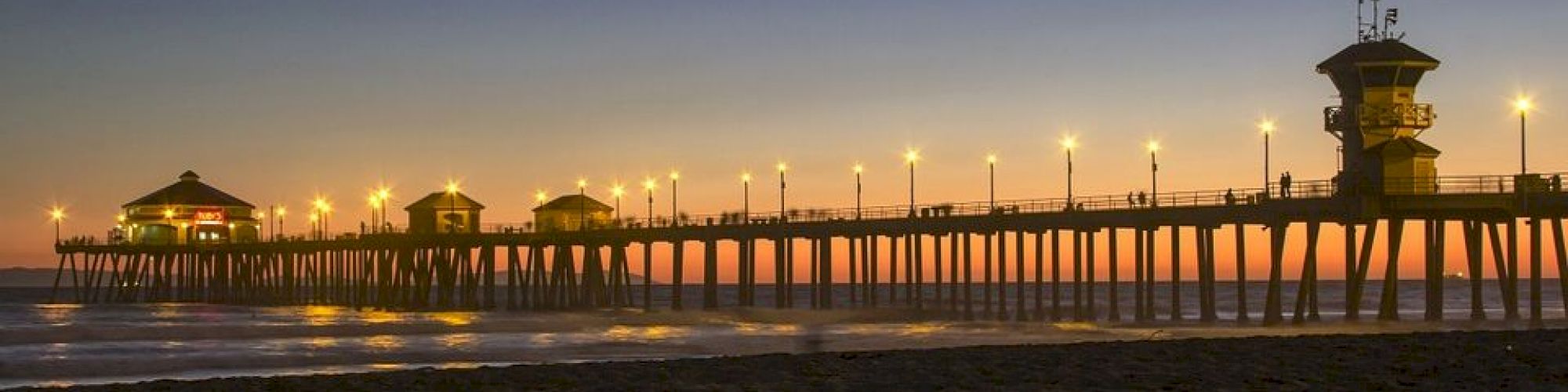 A scenic pier extends over the ocean at sunset, with lights illuminating the surrounding buildings and casting a reflection on the water.