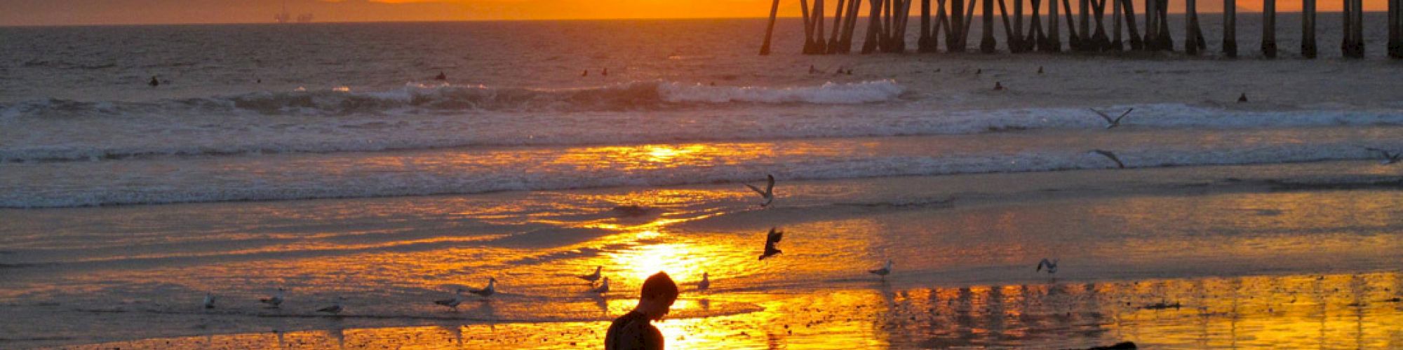 A surfer walks along a beach at sunset with a pier in the background, reflecting the golden hues of the sky in the water.