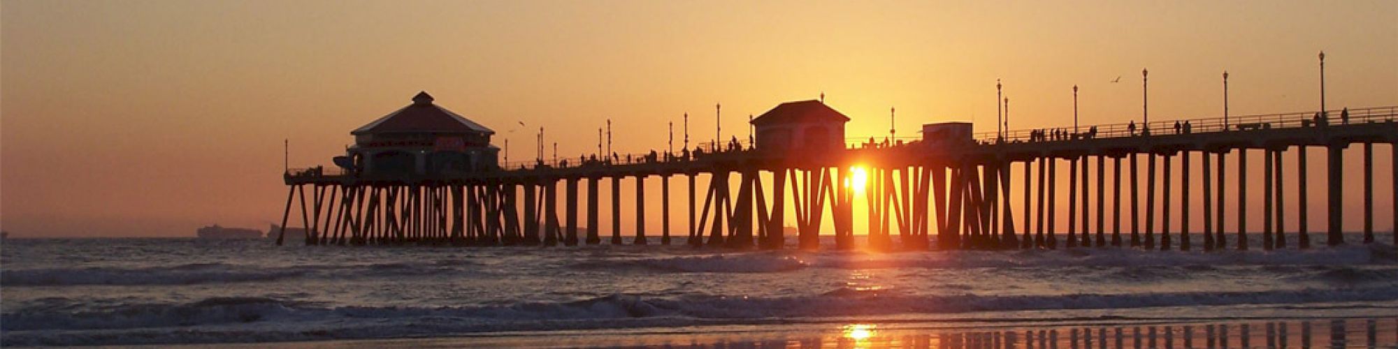 The image shows a pier extending into the ocean with the sun setting behind it, casting a warm glow over the water and sandy beach.