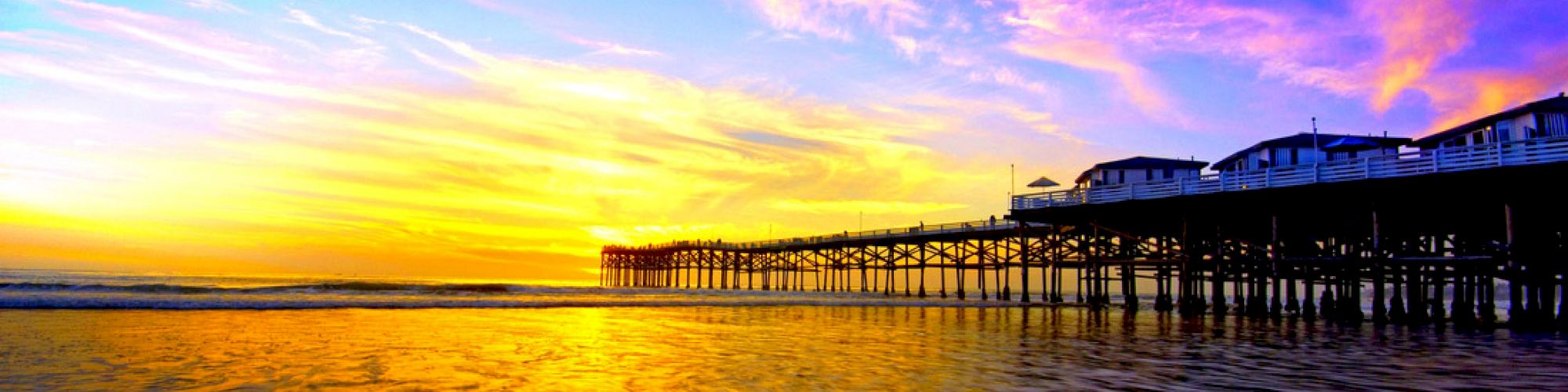 A vibrant sunset over a pier stretching into the ocean, with waves gently washing ashore under a colorful sky of purple, pink, and yellow hues.