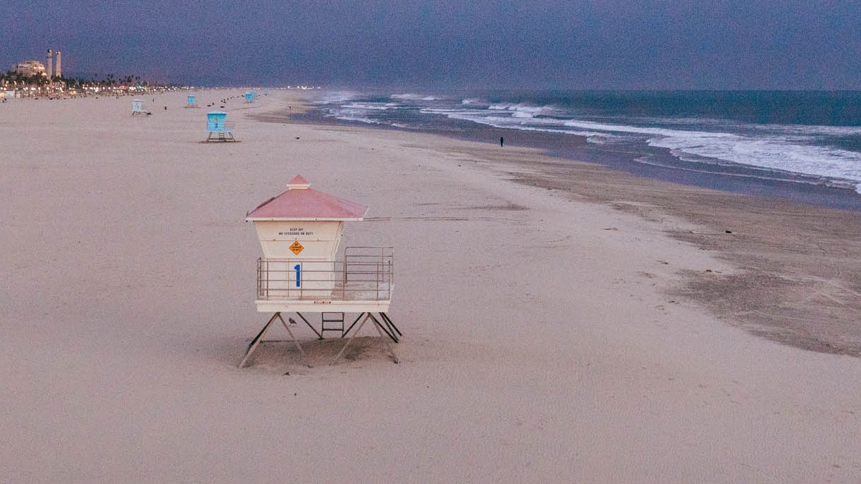 A sandy beach with ocean waves, lifeguard towers, and buildings in the distance, under a cloudy sky. The beach appears deserted.