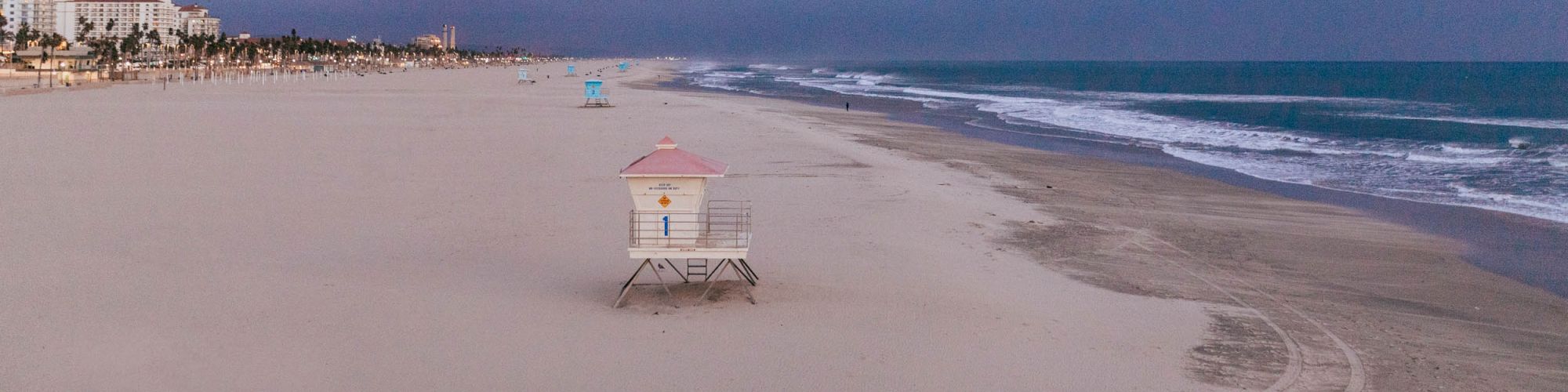 A sandy beach with ocean waves, lifeguard towers, and buildings in the distance, under a cloudy sky. The beach appears deserted.