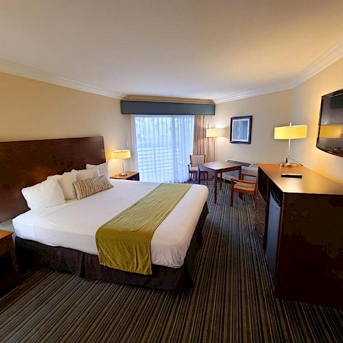 This image shows a neatly arranged hotel room with a king-sized bed, two side tables, a desk, a wall-mounted TV, and a window with curtains.