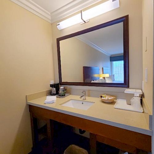 A bathroom sink area with a large mirror, countertop, coffee maker, amenities, and towels, with a waste bin underneath the counter.