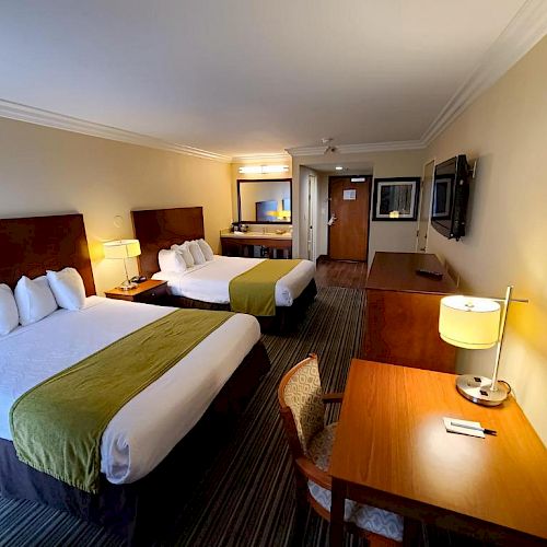 This image shows a hotel room with two beds, a desk with a chair and lamp, a flat-screen TV, and a mirror above a counter in the background.