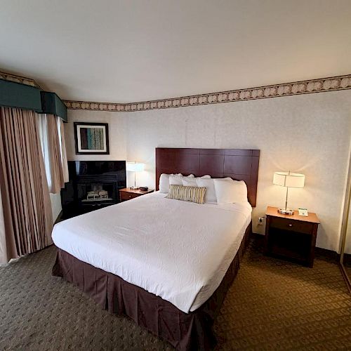 This image shows a hotel room with a large bed, nightstands with lamps, a mirrored closet, and a window with curtains.