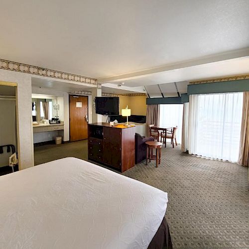 The image shows a spacious hotel room with a bed, TV, dresser, dining table, large windows, and a closet.