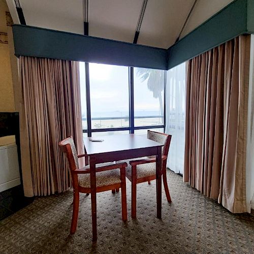 The image shows a small dining area with two wooden chairs and a table by a window with curtains, overlooking a scenic view.