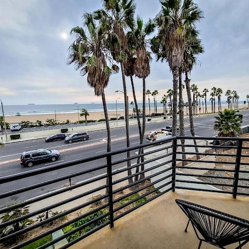 The image shows a coastal view with palm trees lining a road next to a beach. The photo is taken from a balcony with a chair visible.