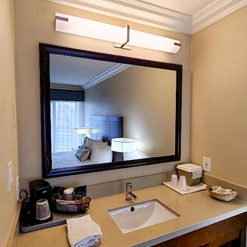 A bathroom vanity area with a large mirror, sink, towels, coffee maker, light fixture above and toiletries arranged neatly on the countertop.