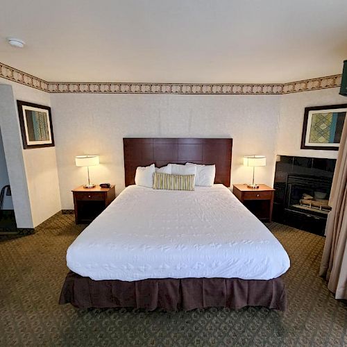 A hotel room with a neatly made bed, two bedside tables with lamps, framed pictures on the walls, and an open closet on the left.