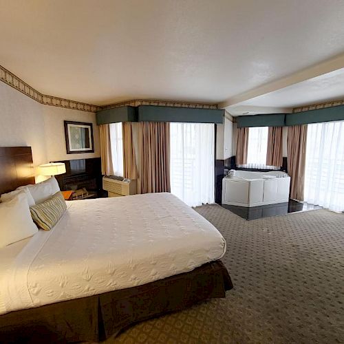 A spacious hotel room with a large bed, white linens, bedside lamps, numerous windows, and a Jacuzzi tub, all exuding comfort and luxury.