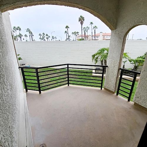 A small balcony with a metal railing, looking out onto a grassy area and palm trees, partly enclosed by stucco walls and an archway.