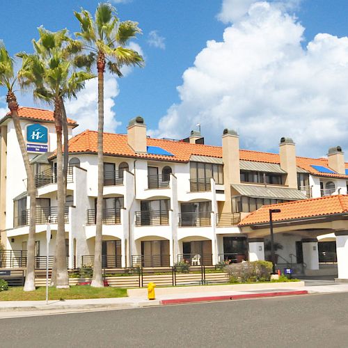 A hotel building with a Mediterranean-style exterior, palm trees, a parking area, and a blue sky with clouds in the background.