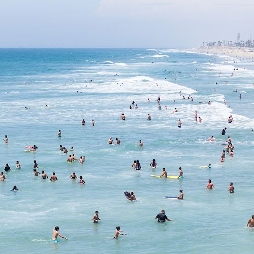 A crowded beach scene with many people swimming and relaxing in the ocean under a clear sky, near a long coastline with visible waves ending the sentence.