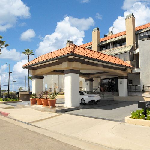 A white car is parked under the entrance canopy of a building with a red-tiled roof, surrounded by palm trees and a clear blue sky.