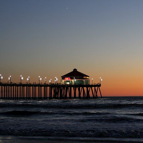 The image shows a pier extending into the ocean at sunset, with lights illuminating the structure and calm waves underneath.