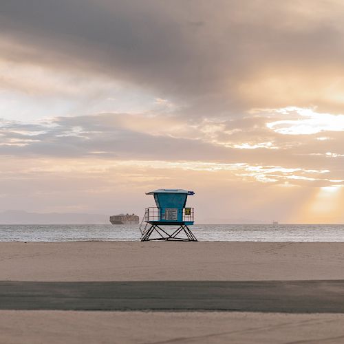 A lifeguard tower stands on a sandy beach with the ocean and ships in the background, under a partly cloudy sunrise sky.