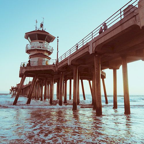 An ocean pier stretches into the sea, with a watchtower and railings above. Waves crash against the supports, and the sky is clear and bright.