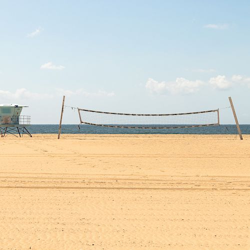 An empty sandy beach with a volleyball net setup and a lifeguard tower in the background, with the ocean and a clear sky beyond.