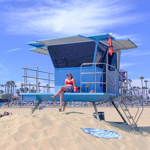 A person sits on a lifeguard tower at a beach, with palm trees and buildings in the background. A blue towel is spread out on the sand below.