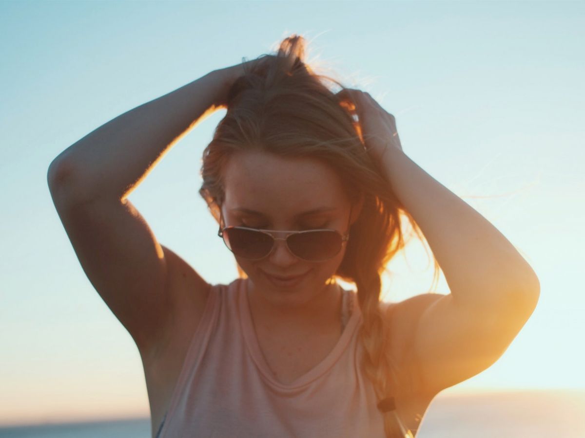 A person wearing sunglasses stands in front of a sunset with hands in their hair, enjoying the moment. The background shows a clear blue sky.