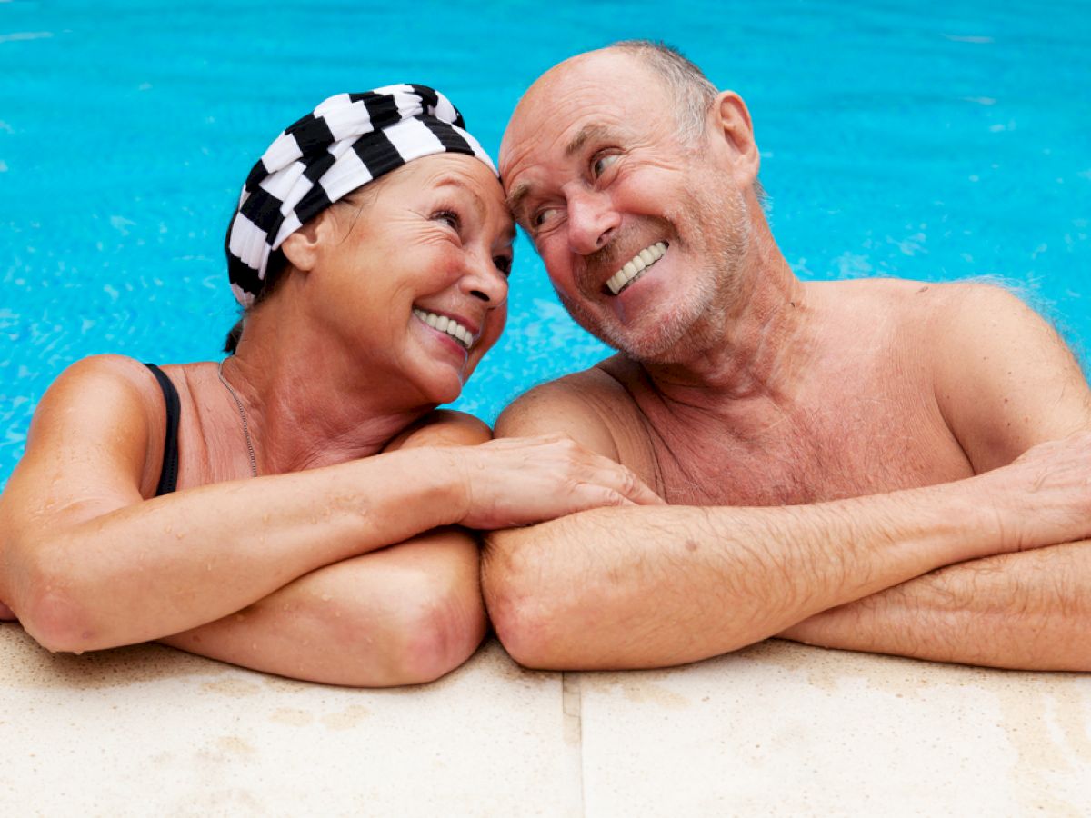 An older couple is smiling and leaning on the edge of a swimming pool, enjoying their time together.