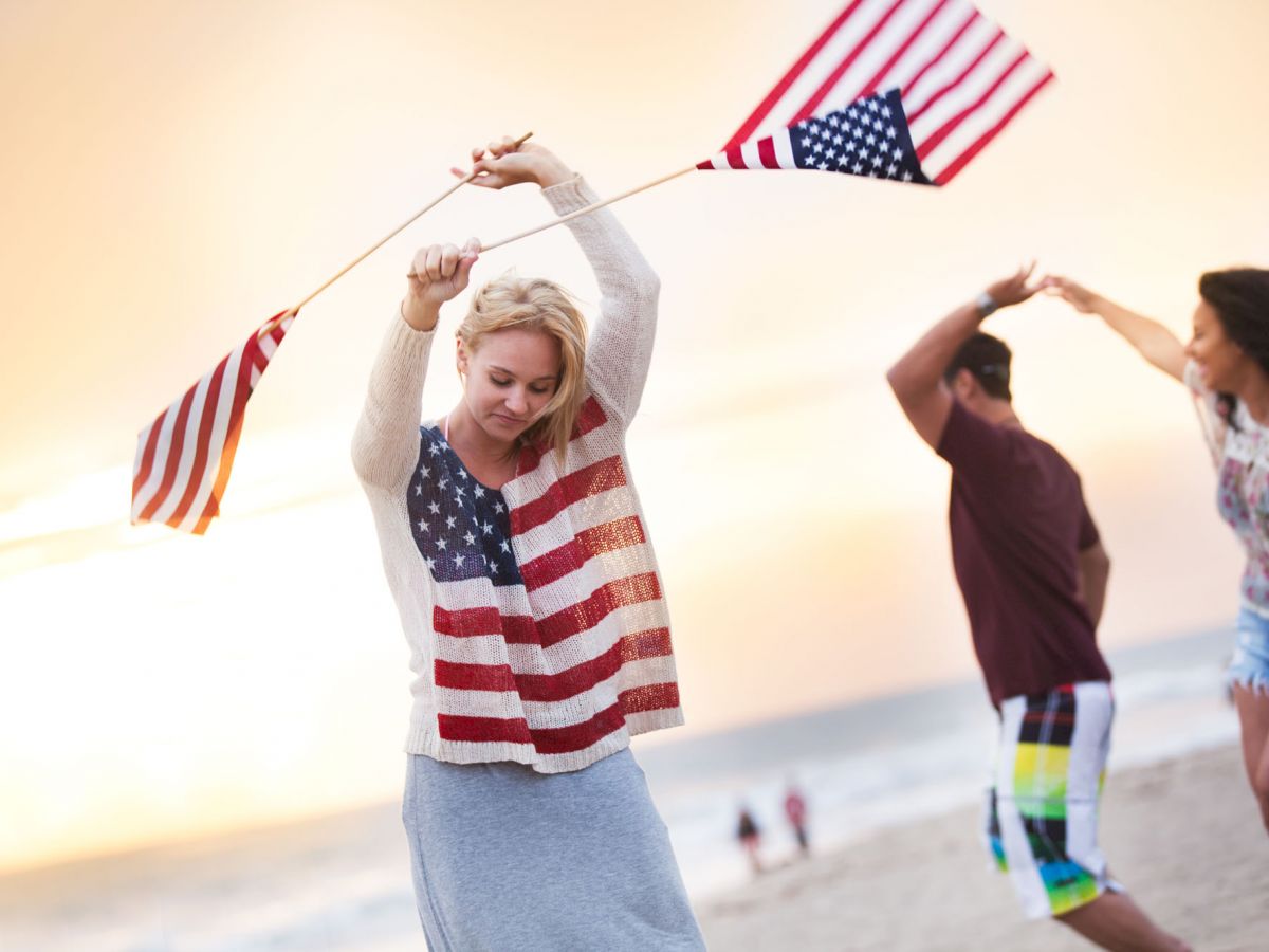 People are on a beach, joyfully waving American flags with a sunset in the background. They appear to be celebrating or having a fun time together.