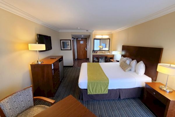 A hotel room with a double bed, nightstands, chairs, desk, lamps, TV, and a dresser. There is a mirror and another lamp on a desk.