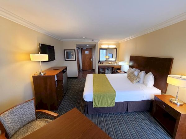 A hotel room with a double bed, nightstands, chairs, desk, lamps, TV, and a dresser. There is a mirror and another lamp on a desk.