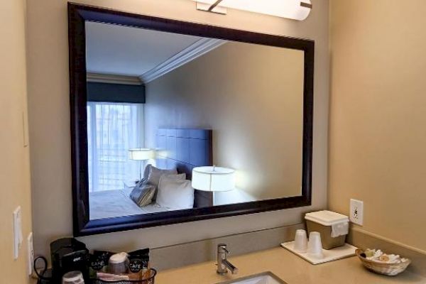 A modern bathroom sink area with a large mirror, light fixture, and countertop with toiletries and a coffee maker on the left side.