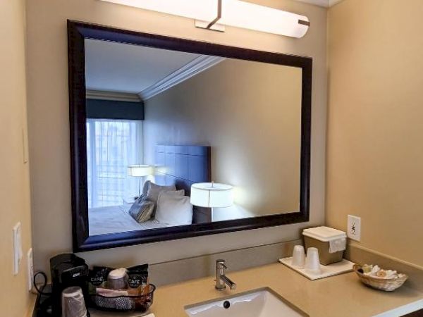 A modern bathroom sink area with a large mirror, light fixture, and countertop with toiletries and a coffee maker on the left side.