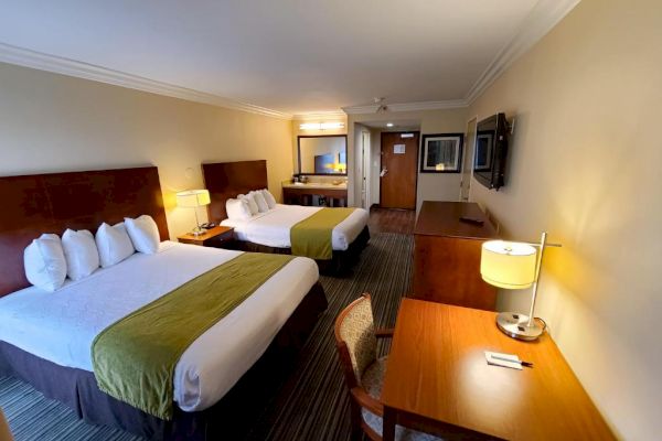 The image shows a hotel room with two beds, a desk, chair, flat-screen TV, and a well-lit mirror near the entrance.