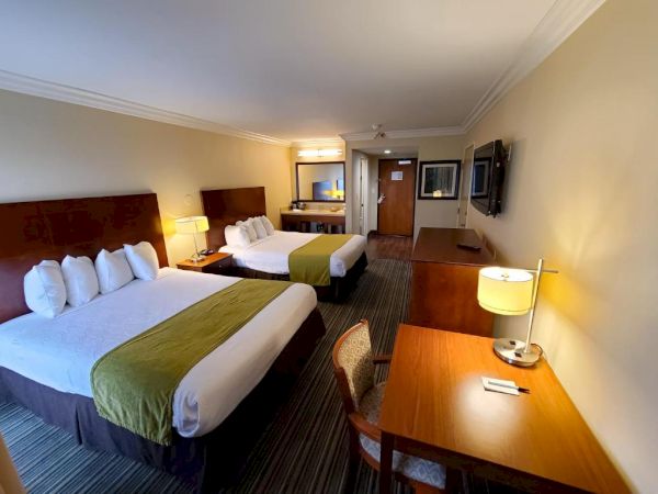 The image shows a hotel room with two beds, a desk, chair, flat-screen TV, and a well-lit mirror near the entrance.