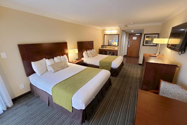 A hotel room features two double beds, a flat-screen TV, a desk, and a chair. There are lamps on nightstands beside the beds and a mirror in the background.