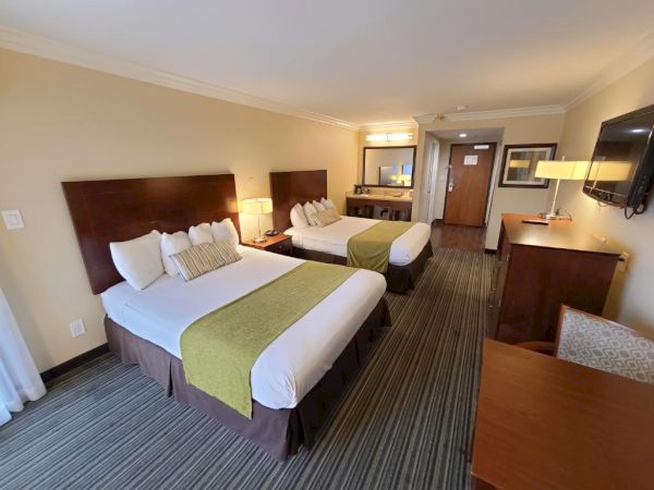 A hotel room features two double beds, a flat-screen TV, a desk, and a chair. There are lamps on nightstands beside the beds and a mirror in the background.