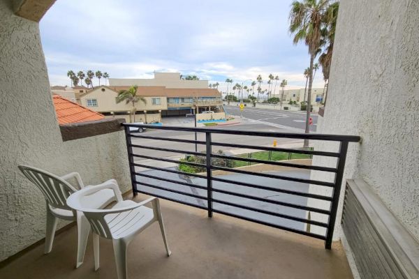 A small balcony with two white plastic chairs overlooks a street with palm trees, buildings, and a cloudy sky in the background.