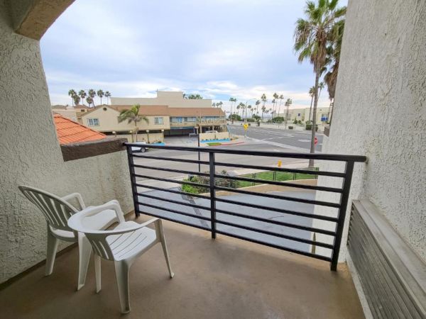 A small balcony with two white plastic chairs overlooks a street with palm trees, buildings, and a cloudy sky in the background.