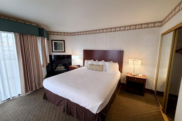 A hotel room with a large bed, nightstands with lamps, a dresser, and a closet with mirrored doors. Wall art and curtains add decor.