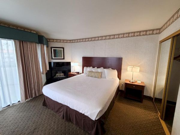 A hotel room with a large bed, nightstands with lamps, a dresser, and a closet with mirrored doors. Wall art and curtains add decor.