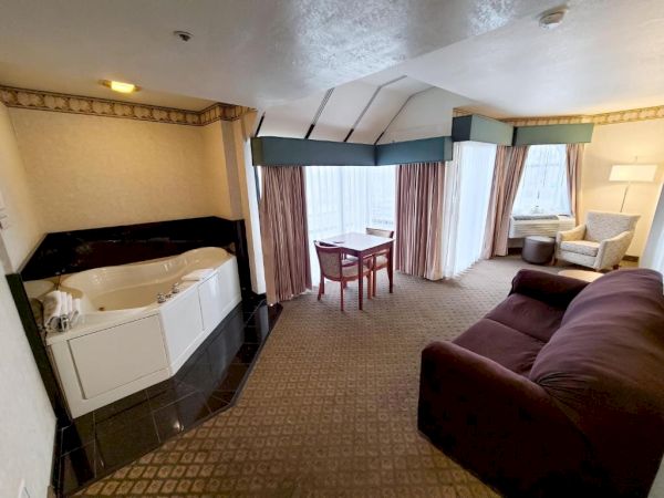 A cozy hotel room features a jacuzzi tub, a small dining table with two chairs, a couch, and an armchair with a floor lamp, all near large windows.