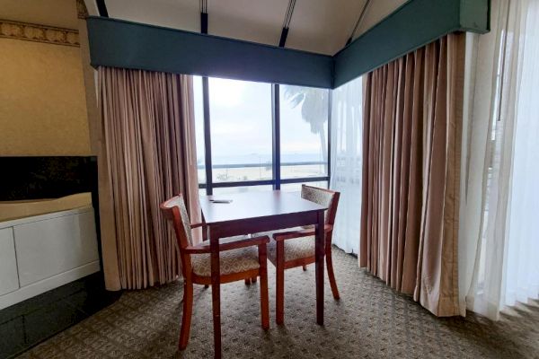 The image shows a small dining area with a table and two chairs facing a large window covered by curtains, offering a view of the outside.