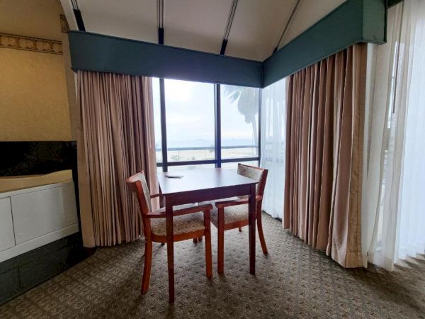 The image shows a small dining area with a table and two chairs facing a large window covered by curtains, offering a view of the outside.